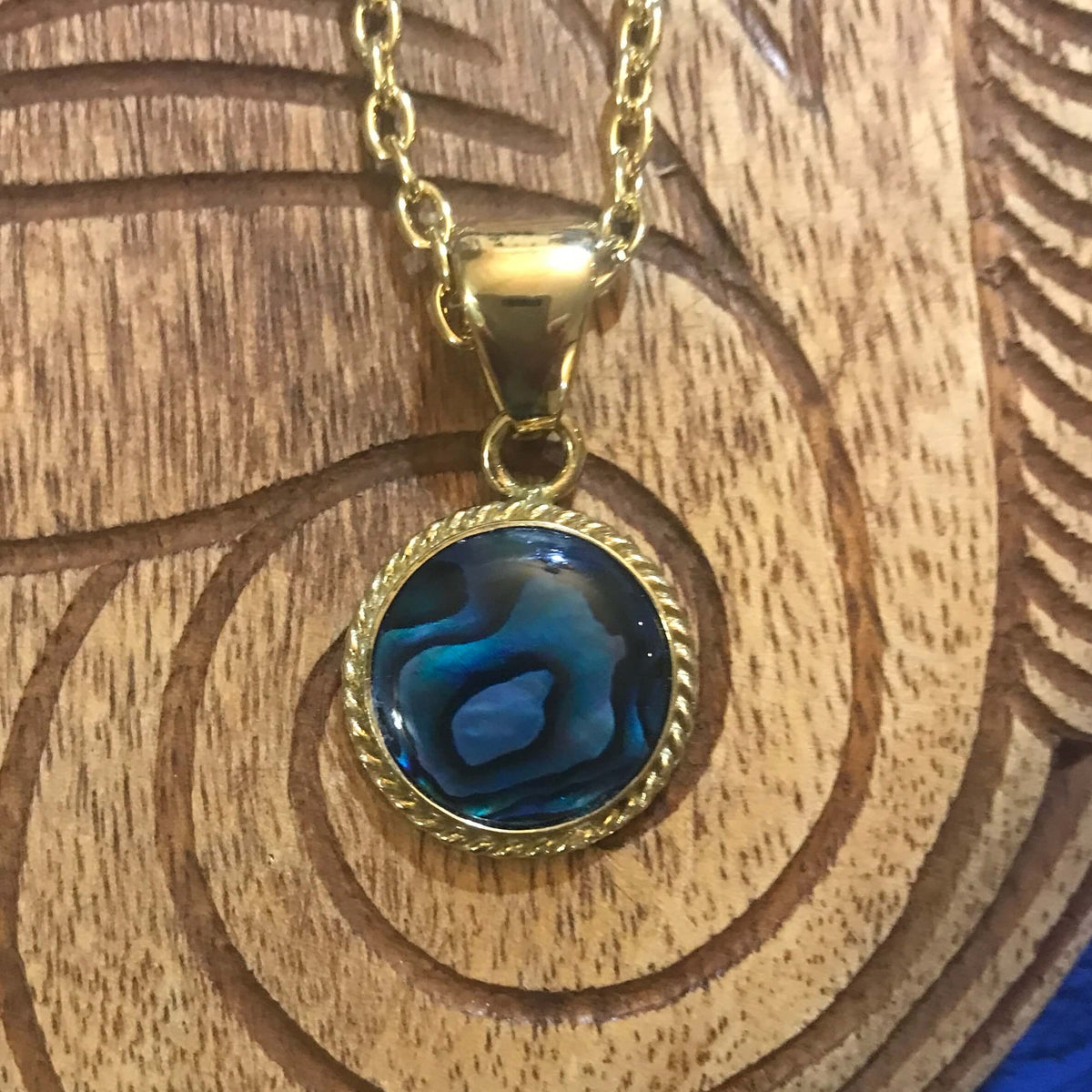 Close up view of a round blue abalone pendant with alchemia gold rope trim around the shell