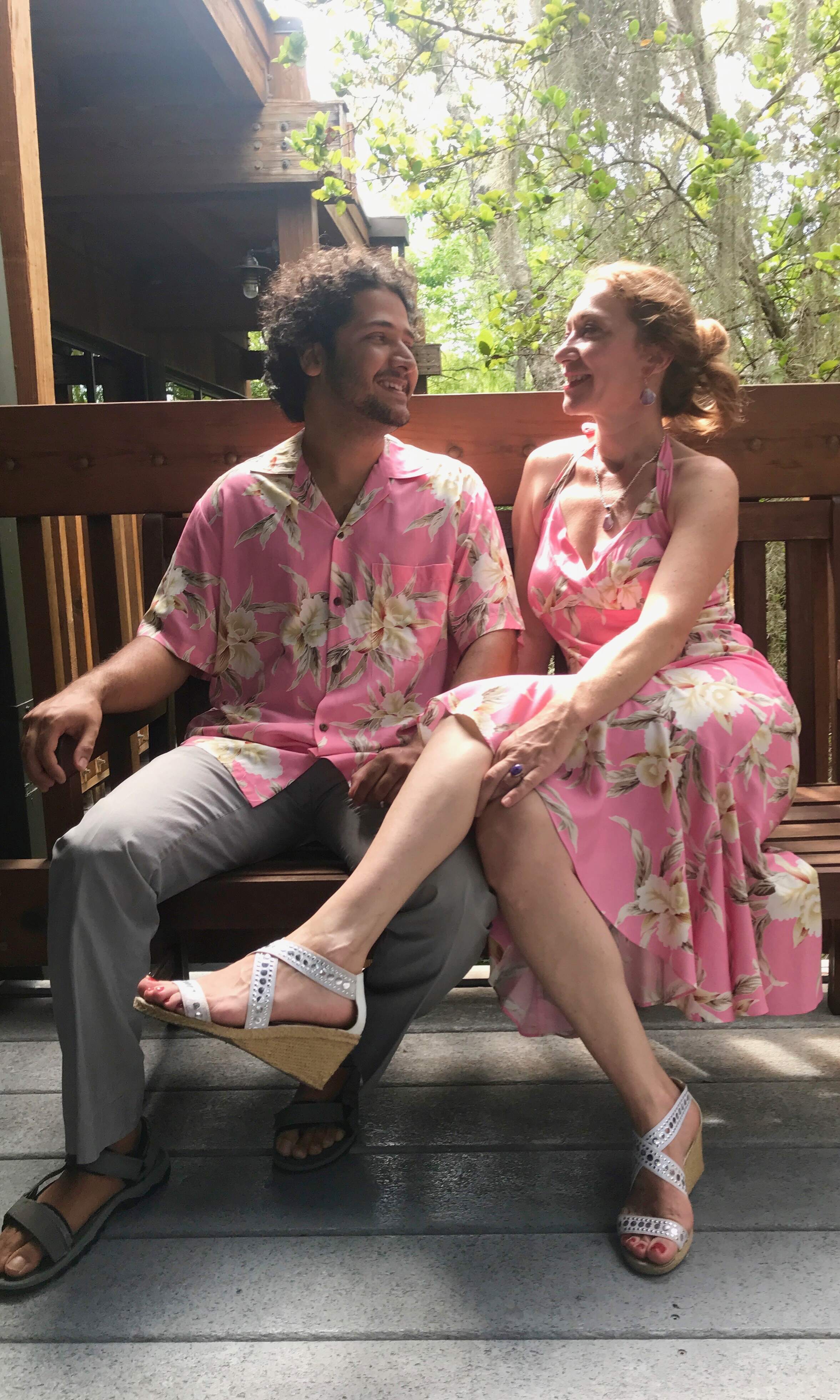 Hawaiian couple wearing matching pink orchid dress and shirt outfits sitting on a bench together | Aloha Products USA